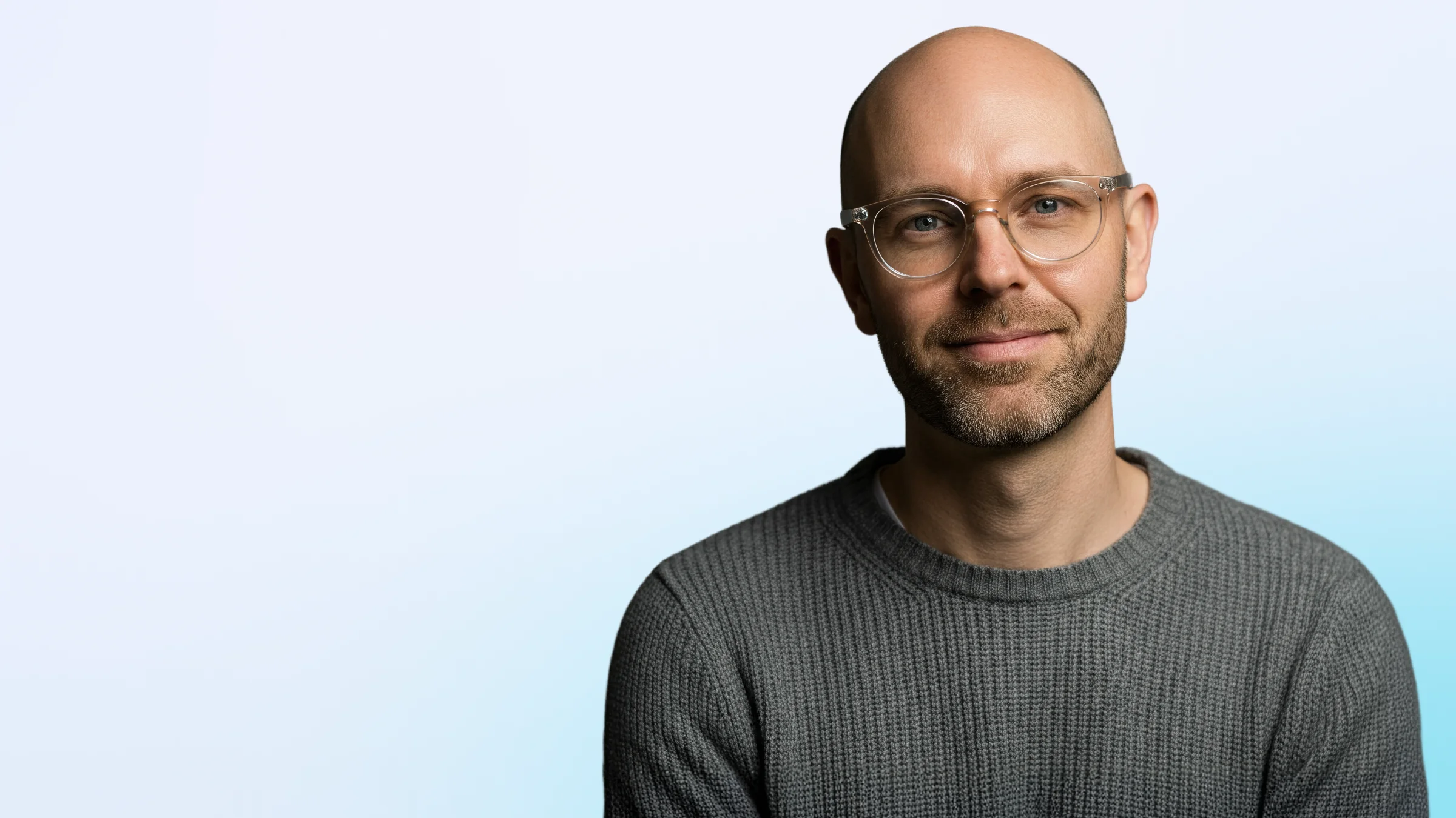 Smiling bald man with glasses wearing a gray sweater against a light blue background.