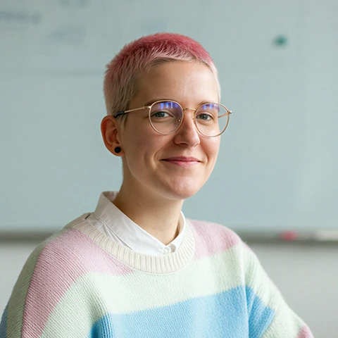 Young adult with short pink hair, glasses, and a pastel striped sweater, smiling and sitting in front of a blurred whiteboard.